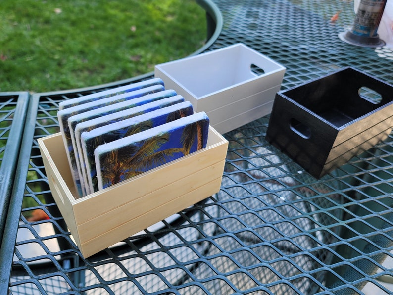 Puede incluir: Un juego de posavasos rectangulares con un dise&ntilde;o de palmera y cielo azul, exhibidos en una caja de madera clara. Tambi&eacute;n se ven dos cajas de madera adicionales, una blanca y otra negra.