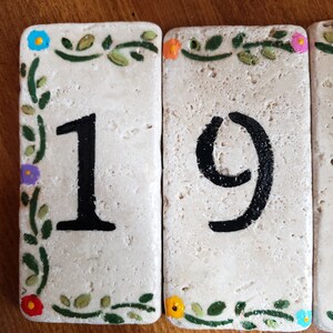 May include: Two white ceramic tiles with green vines and colorful flowers painted around the edges. The tiles have black painted numbers, "19", on them.