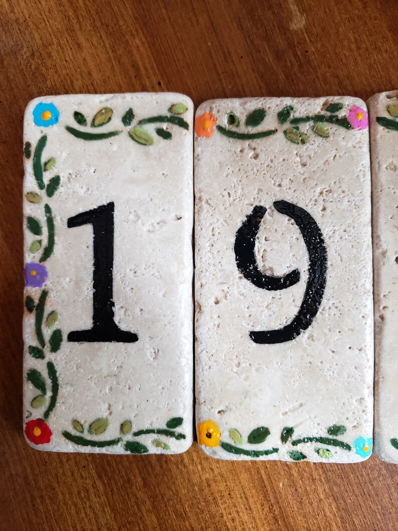 May include: Two white ceramic tiles with green vines and colorful flowers painted around the edges. The tiles have black painted numbers, "19", on them.