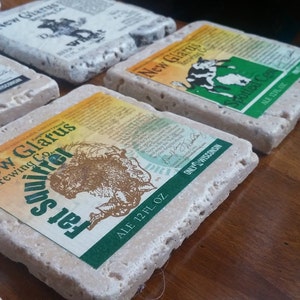 May include: Six square stone coasters with a light brown colour and a rough texture. Each coaster features a different design with a New Glarus Brewing Co. logo and a beer name. The designs include a squirrel, a cow, and a paw print.