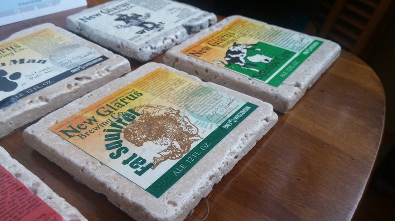 May include: Six square stone coasters with a light brown colour and a rough texture. Each coaster features a different design with a New Glarus Brewing Co. logo and a beer name. The designs include a squirrel, a cow, and a paw print.