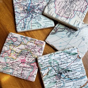 May include: A set of five square stone coasters featuring vintage map prints of various cities including Budapest, Moscow, Sochi, Frankfurt, and Wien. Each coaster displays detailed cartographic lines, city names, and geographic labels.