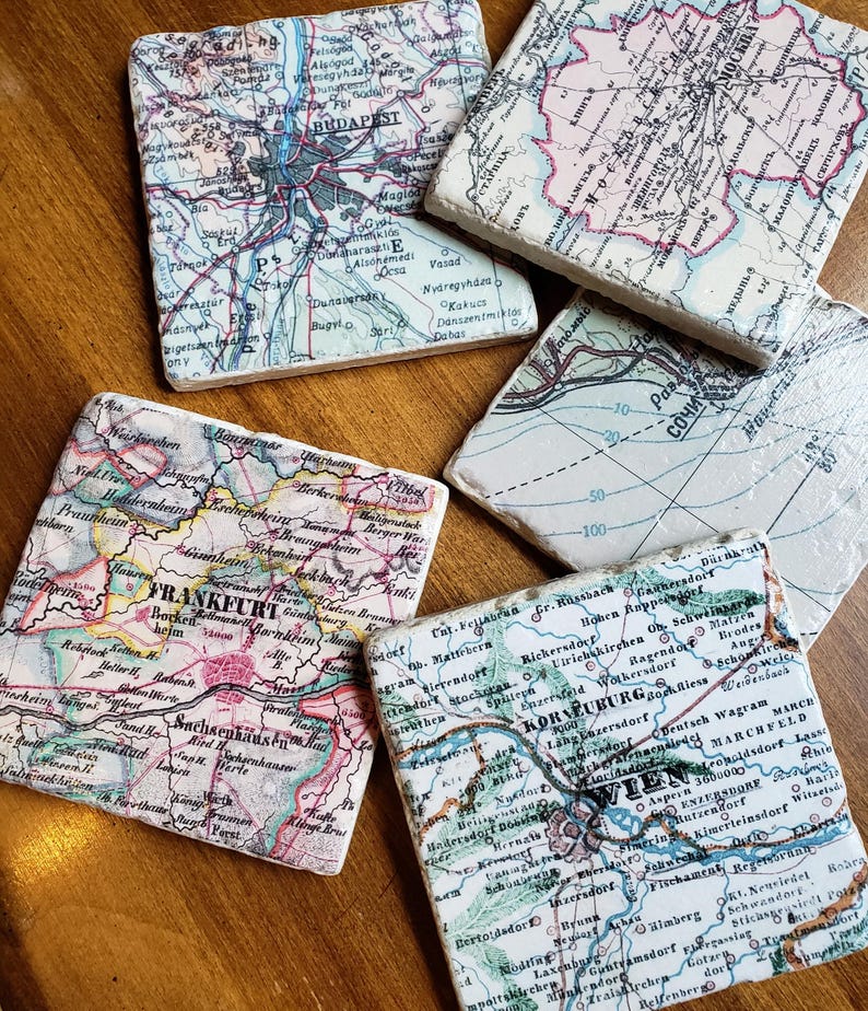 May include: A set of five square stone coasters featuring vintage map prints of various cities including Budapest, Moscow, Sochi, Frankfurt, and Wien. Each coaster displays detailed cartographic lines, city names, and geographic labels.