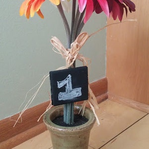 May include: A bouquet of pink, orange, and purple flowers in a green ceramic vase with a small black chalkboard sign that says "1" on it.
