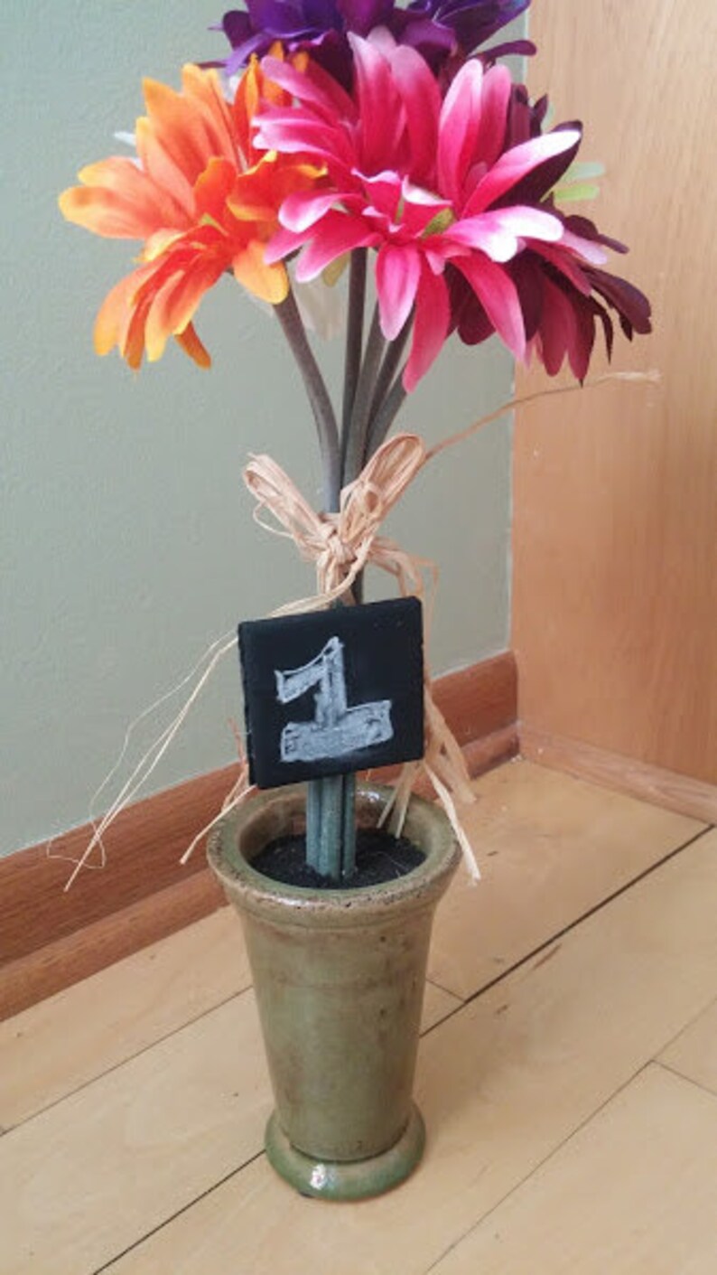May include: A bouquet of pink, orange, and purple flowers in a green ceramic vase with a small black chalkboard sign that says "1" on it.