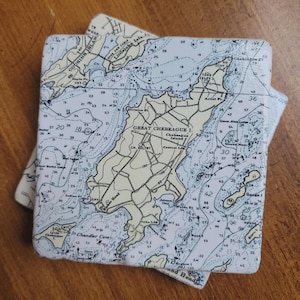 May include: Two square coasters with a nautical chart design. The charts feature islands, depth soundings, and place names in blue and beige. The coasters are on a wooden surface.