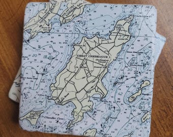 May include: Two square coasters with a nautical chart design. The charts feature islands, depth soundings, and place names in blue and beige. The coasters are on a wooden surface.