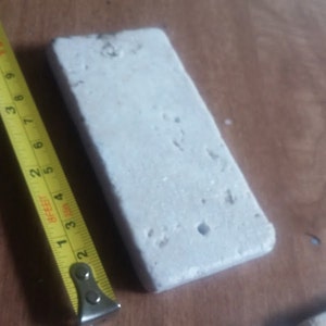 May include: A white rectangular tile with a rough texture and small holes. The tile is being measured with a yellow tape measure.