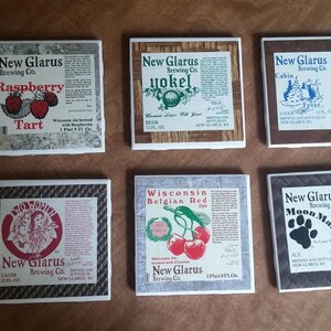 May include: Six square ceramic coasters featuring different New Glarus Brewing Co. beers. The coasters feature images of the beers, including Raspberry Tart, Yokel, Cabin Fever, Wisconsin Belgian Red, and Moon Man.