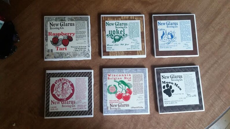 May include: Six square ceramic coasters featuring different New Glarus Brewing Co. beers. The coasters feature images of the beers, including Raspberry Tart, Yokel, Cabin Fever, Wisconsin Belgian Red, and Moon Man.