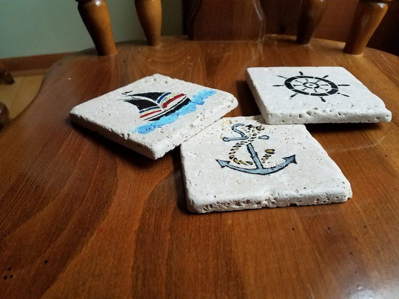May include: Three square stone coasters with nautical designs. One coaster features a blue and white sailboat, another has a black anchor with a gold rope, and the third has a black steering wheel.