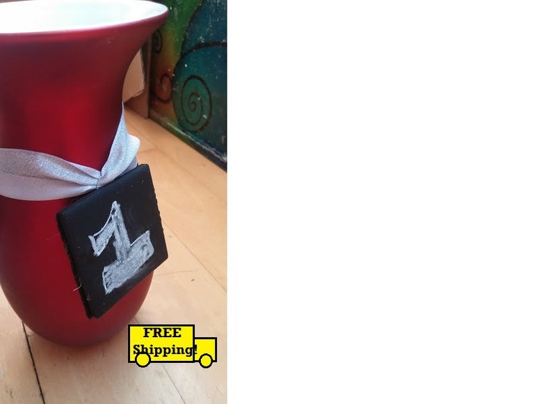 May include: A red ceramic vase with a white ribbon tied around the neck. A small black chalkboard tag with the number "1" written in white chalk is attached to the ribbon.