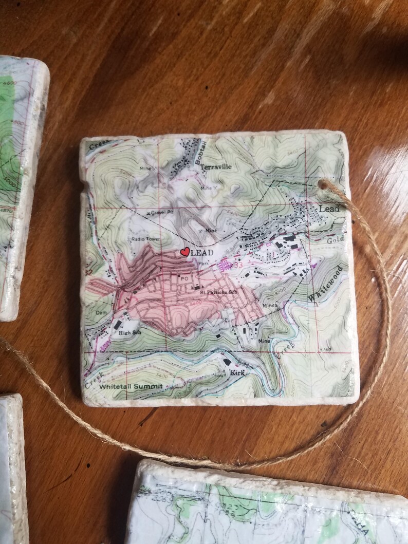 K&ouml;nnte beinhalten: Eine wei&szlig;e Keramikfliese mit einer Vintage-Karte von Lead, South Dakota. Die Karte ist in Braun, Gr&uuml;n und Rosa gedruckt. Ein rotes Herz ist &uuml;ber die Stadt Lead gelegt. Die Fliese hat ein Loch in der oberen Ecke und eine braune Schnur daran befestigt.