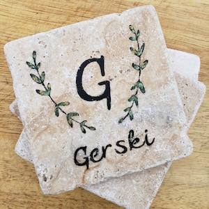May include: Set of four beige stone coasters with a green leafy design and the letter "G" and the name "Gerski" written in black.