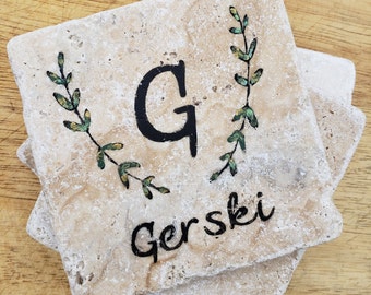 May include: Set of four beige stone coasters with a green leafy design and the letter "G" and the name "Gerski" written in black.