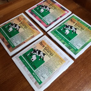 May include: Four white square coasters with a green, black, and white design featuring a spotted cow and the text "New Glarus Brewing Co. Spotted Cow Ale 12 fl oz. Only in Wisconsin."