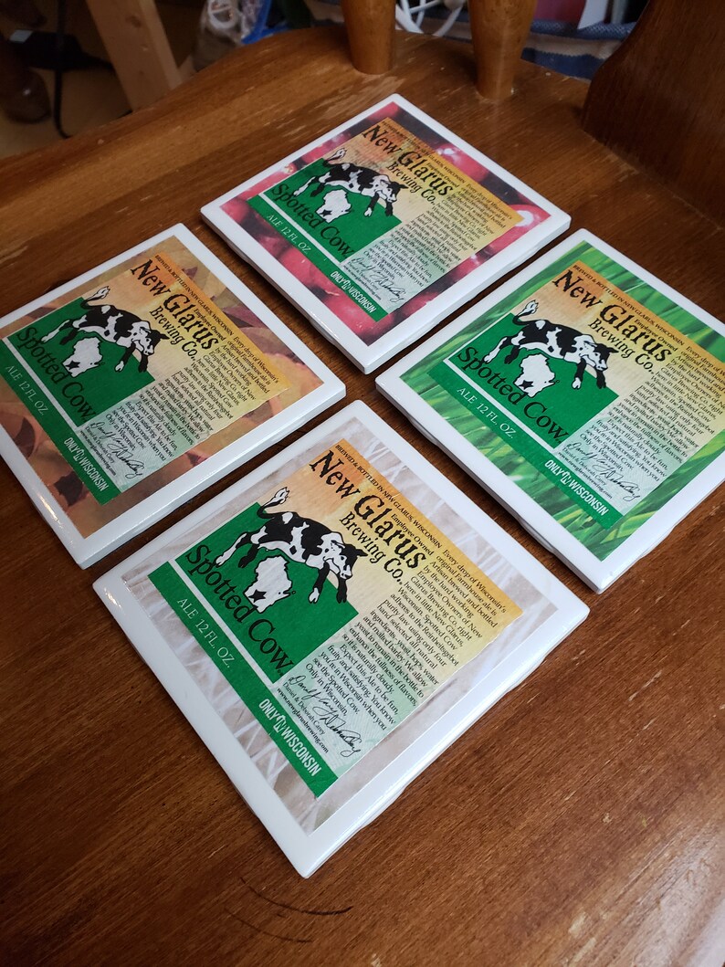 May include: Four white square coasters with a green, black, and white design featuring a spotted cow and the text "New Glarus Brewing Co. Spotted Cow Ale 12 fl oz. Only in Wisconsin."
