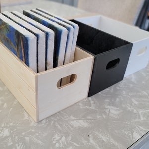 Puede incluir: Una caja de madera llena de posavasos rectangulares con una escena de palmeras tropicales. Junto a ella hay dos cajas rectangulares, una negra y otra blanca, cada una con un recorte de asa. Los art&iacute;culos est&aacute;n sobre una superficie gris claro.