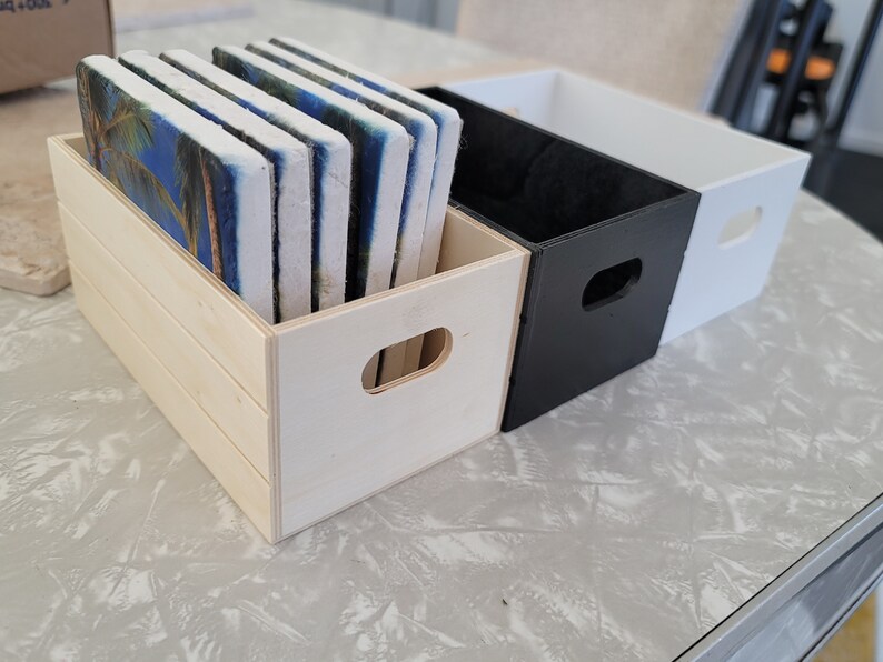Puede incluir: Una caja de madera llena de posavasos rectangulares con una escena de palmeras tropicales. Junto a ella hay dos cajas rectangulares, una negra y otra blanca, cada una con un recorte de asa. Los art&iacute;culos est&aacute;n sobre una superficie gris claro.