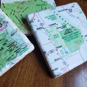 May include: Four square coasters with map designs. The coasters feature sections of maps with green, white, and tan colors. The maps show geographical features, roads, and place names. The coasters are on a wooden surface.