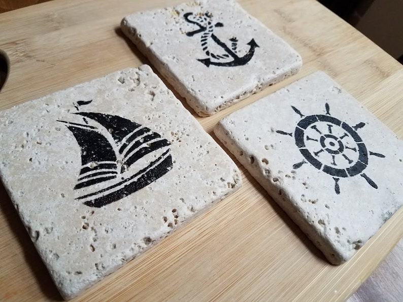 May include: Set of three square stone coasters with nautical designs. The coasters feature a sailboat, an anchor, and a ship's wheel.