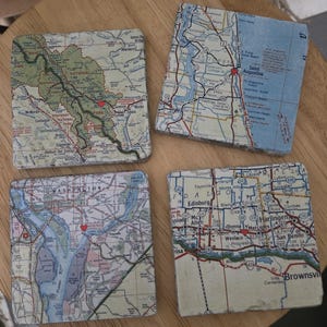 May include: Four square stone coasters featuring printed map sections of various regions, including Banff National Park, Saint Augustine, Washington, and the Brownsville area, each marked with a small red heart icon at specific locations.
