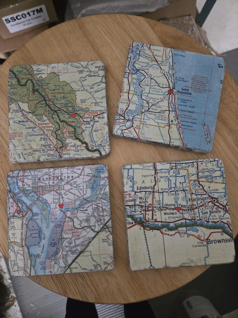 May include: Four square stone coasters featuring printed map sections of various regions, including Banff National Park, Saint Augustine, Washington, and the Brownsville area, each marked with a small red heart icon at specific locations.