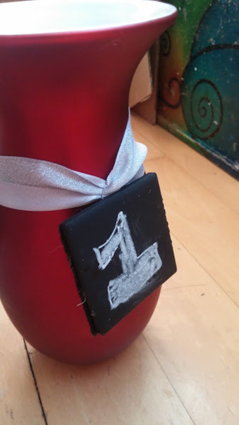 May include: A red ceramic vase with a white ribbon tied around it. A black tag with the number "1" written in white chalk is attached to the ribbon.