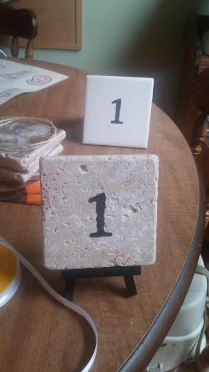 May include: A beige tile with the number "1" painted in black on it. The tile is on a small black easel.