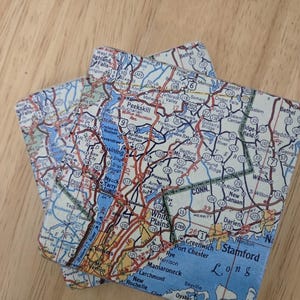 May include: Two square coasters featuring a detailed map print of the Westchester County and Hudson Valley region in New York. The map displays various towns, highways, and water bodies in shades of blue, red, yellow, and white with black text.