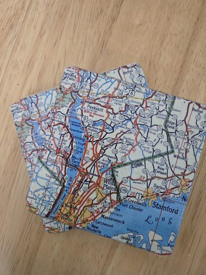 May include: Two square coasters featuring a detailed map print of the Westchester County and Hudson Valley region in New York. The map displays various towns, highways, and water bodies in shades of blue, red, yellow, and white with black text.