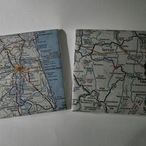 May include: Two square coasters featuring printed map designs. The left coaster displays a map of the Jacksonville area with a small red heart icon. The right coaster shows a map of the Bellows Falls region with a small red heart icon.