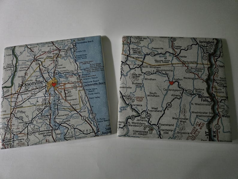 May include: Two square coasters featuring printed map designs. The left coaster displays a map of the Jacksonville area with a small red heart icon. The right coaster shows a map of the Bellows Falls region with a small red heart icon.