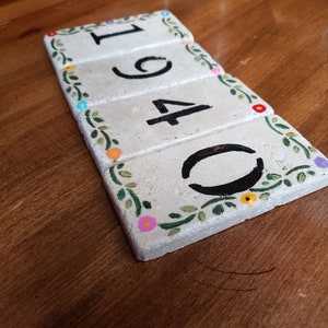 May include: A set of four white ceramic tiles with black numbers 1940 and a floral design around the edges. The tiles are arranged in a row and have a rustic, handmade look.