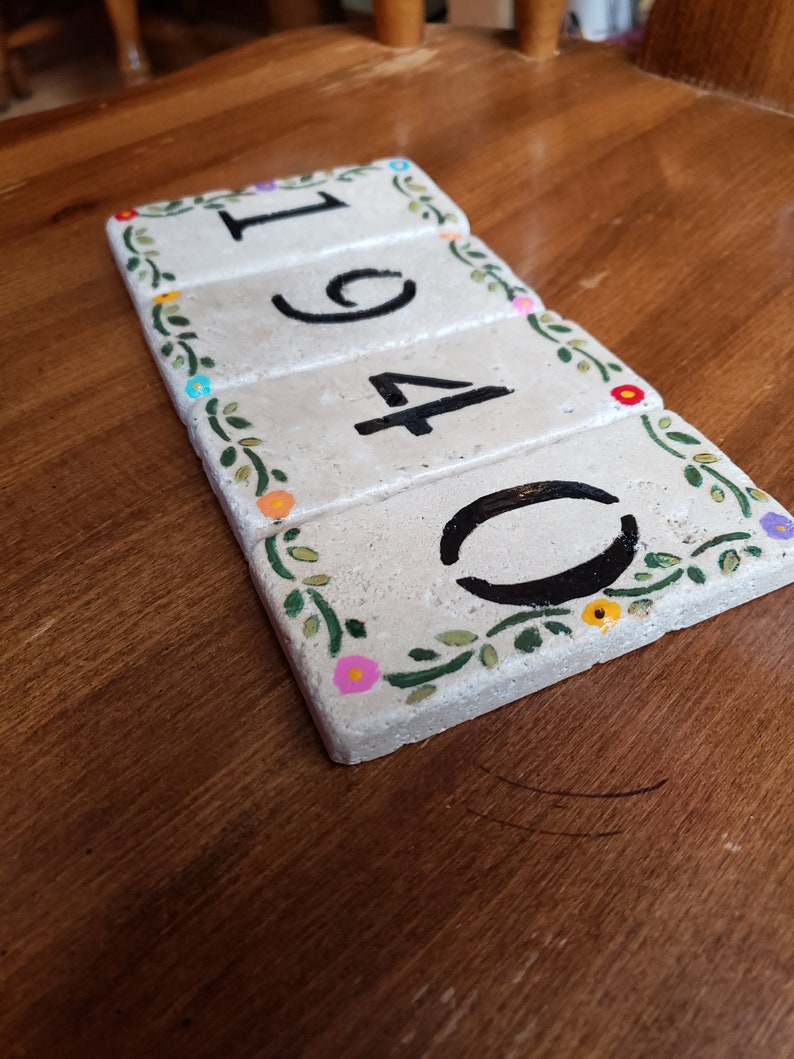 May include: A set of four white ceramic tiles with black numbers 1940 and a floral design around the edges. The tiles are arranged in a row and have a rustic, handmade look.