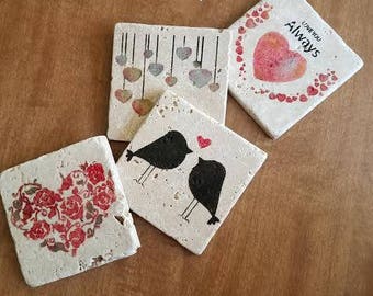 May include: Four white ceramic coasters with different designs. One coaster features a heart with the words "Love You Always". Another coaster has a design of hanging hearts. A third coaster has a design of two birds. The final coaster has a design of a heart made of smaller hearts.