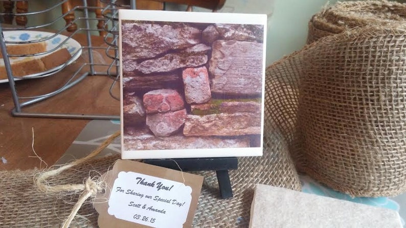 Puede incluir: Un peque&ntilde;o azulejo de cer&aacute;mica blanco con una fotograf&iacute;a de un muro de piedra. El muro est&aacute; hecho de piedras de varios tama&ntilde;os en tonos marrones, grises y rojos. El azulejo est&aacute; sobre un peque&ntilde;o caballete con una etiqueta de agradecimiento que dice "&iexcl;Gracias! Por compartir nuestro d&iacute;a especial! Scott & Amanda 03.06.15".