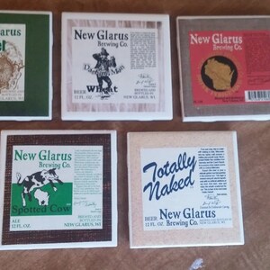 May include: Five square ceramic coasters with different designs featuring New Glarus Brewing Co. beer labels. The coasters feature images of a squirrel, a man in a hat, a map of Wisconsin, a cow, and the words "Totally Naked".