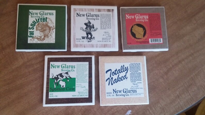 May include: Five square ceramic coasters with different designs featuring New Glarus Brewing Co. beer labels. The coasters feature images of a squirrel, a man in a hat, a map of Wisconsin, a cow, and the words "Totally Naked".