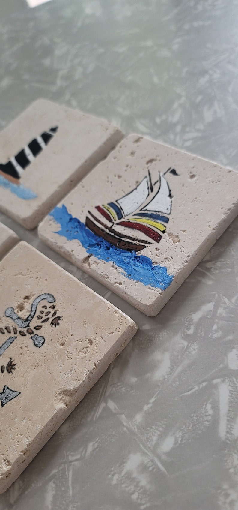 May include: Set of three square stone coasters with nautical themed designs. The coasters feature a lighthouse, a sailboat, and an anchor. The coasters are painted in black, white, blue, red, and yellow.