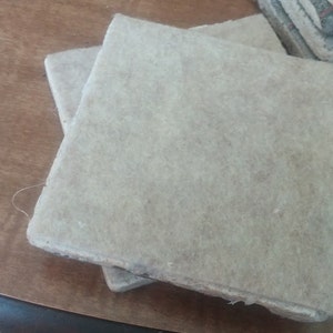 Puede incluir: Primer plano de varios posavasos cuadrados de color claro. Los posavasos parecen estar hechos de un material fibroso, posiblemente papel o una sustancia similar. Est&aacute;n apilados sobre una superficie de madera marr&oacute;n.