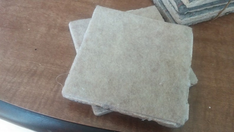Puede incluir: Primer plano de varios posavasos cuadrados de color claro. Los posavasos parecen estar hechos de un material fibroso, posiblemente papel o una sustancia similar. Est&aacute;n apilados sobre una superficie de madera marr&oacute;n.