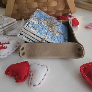 May include: A stack of square ceramic coasters featuring a blue and white map print, tied with brown twine, resting inside a tan leather tray with brass-coloured metal rivets at the corners, surrounded by small red and white felt heart shapes.