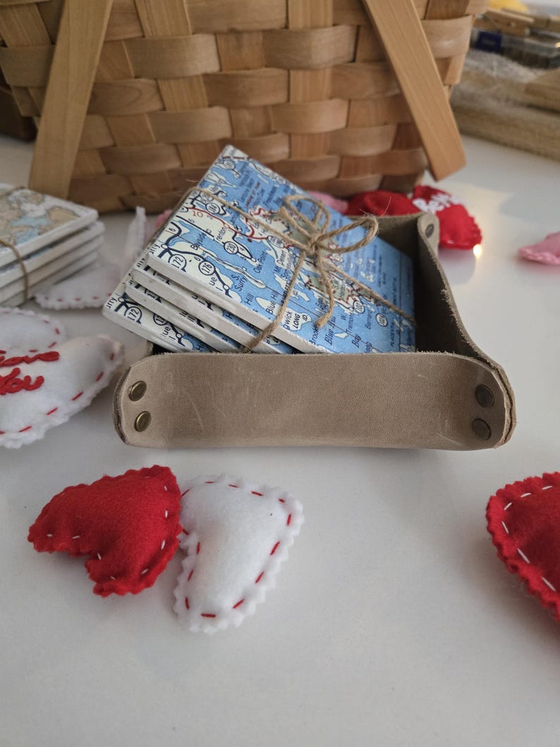 May include: A stack of square ceramic coasters featuring a blue and white map print, tied with brown twine, resting inside a tan leather tray with brass-coloured metal rivets at the corners, surrounded by small red and white felt heart shapes.