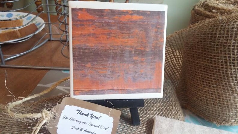 Puede incluir: Un posavasos de madera r&uacute;stico con un acabado naranja y marr&oacute;n desgastado. El posavasos est&aacute; en un peque&ntilde;o caballete con una etiqueta de agradecimiento que dice "&iexcl;Gracias! Por compartir nuestro d&iacute;a especial! Scott & Amanda".