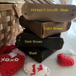 May include: A stack of four square leather valet trays in khaki, light brown, dark brown, and black, each secured with metal rivets at the corners. The trays are labelled with text indicating their colours, surrounded by small felt heart decorations.