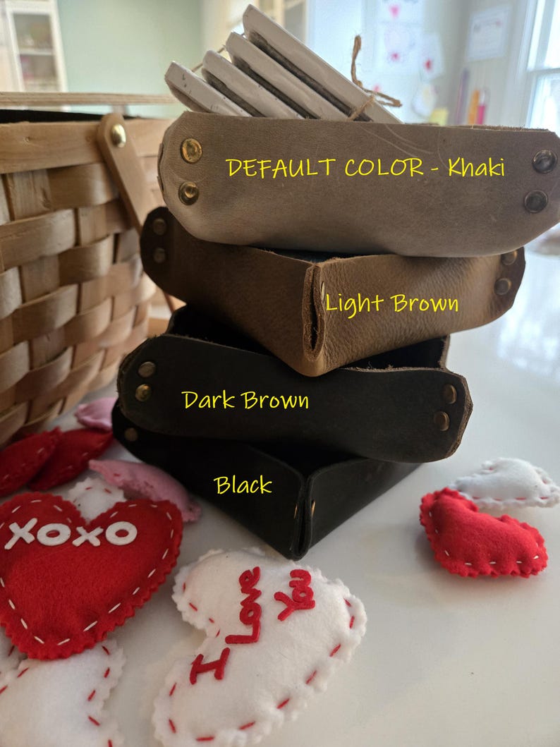 May include: A stack of four square leather valet trays in khaki, light brown, dark brown, and black, each secured with metal rivets at the corners. The trays are labelled with text indicating their colours, surrounded by small felt heart decorations.
