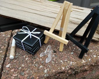 May include: A stack of black square coasters with a white ribbon tied around them. The coasters are sitting on a wooden surface next to two small wooden easels, one black and one natural wood.