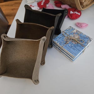 May include: Three square leather valet trays in tan, brown, and black, each featuring metal snap fasteners at the corners. Beside the trays sits a stack of square coasters with a blue map print tied with twine, and small felt heart decorations.