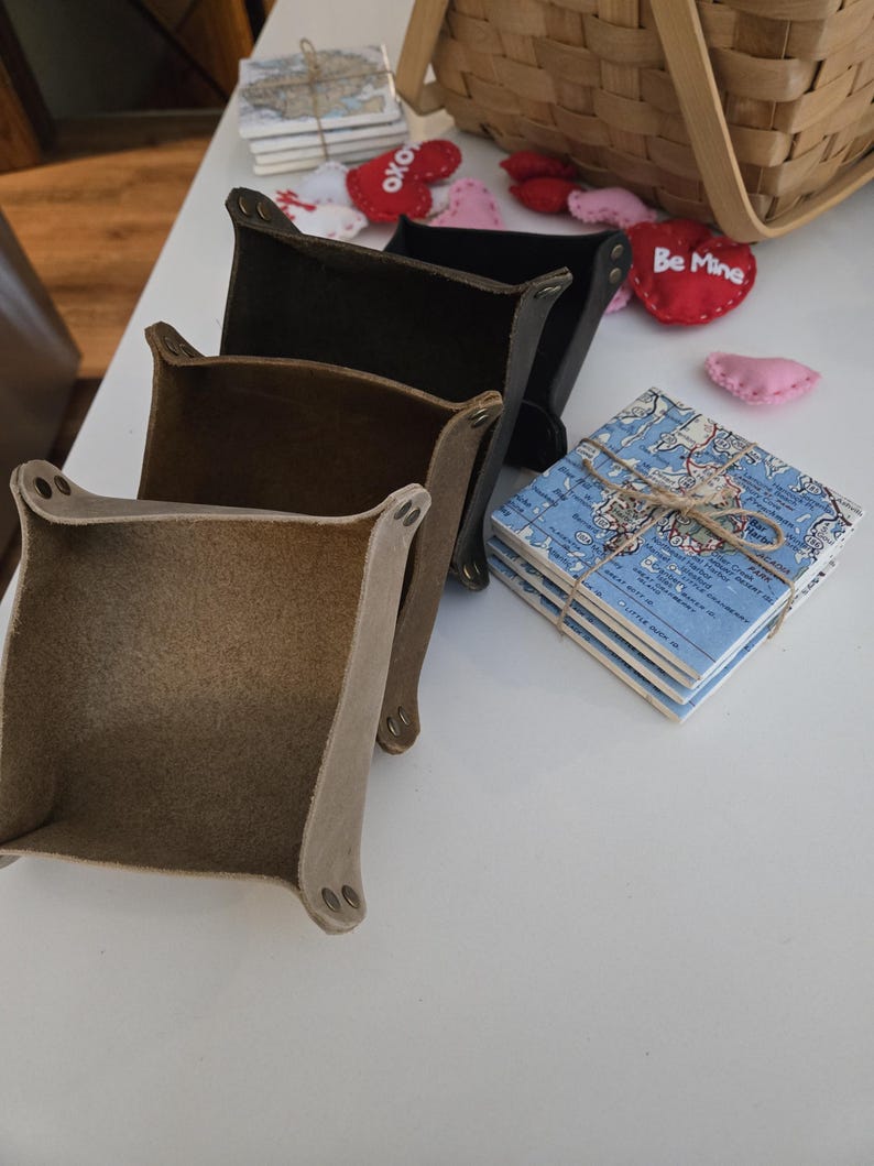 May include: Three square leather valet trays in tan, brown, and black, each featuring metal snap fasteners at the corners. Beside the trays sits a stack of square coasters with a blue map print tied with twine, and small felt heart decorations.
